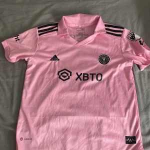 Inter Miami Messi 10 rosa fotbollströja från Adidas - Säljer en rosa Inter Miami fotbollströja med Messi 10-tryck. Tröjan har svarta detaljer, klubbmärke på bröstet och flera sponsorer. Klassisk krage och tre svarta ränder på axlarna. Perfekt för fans av både laget och spelaren.