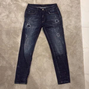 Mörkblå jeans med slitningar från Alessandro Zavetti - Säljer ett par mörkblå jeans från Alessandro Zavetti med coola slitningar och färgstänk. Byxorna har klassisk femficksdesign, knappgylf och raka ben. Perfekta för dig som vill ha en trendig och avslappnad stil.