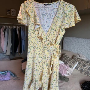 Gul blommig omlottklänning från SHEIN - Söt gul omlottklänning med blommigt mönster och volangdetaljer. Klänningen har korta ärmar och knyt i midjan. 