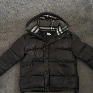 Svart pufferjacka från Burberry - Säljer en svart pufferjacka från Burberry med huva och klassiskt rutigt foder. Jackan har dragkedja och knappar framtill samt två fickor med dragkedja. Perfekt för kyliga dagar och riktigt snygg streetstil.