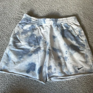 Nike batikmönstrade shorts - Säljer ett par vita och ljusblå batikmönstrade shorts från Nike. De har resår i midjan och sidofickor, samt en liten svart Nike-logga på ena benet. Perfekta för en avslappnad stil under varma dagar.