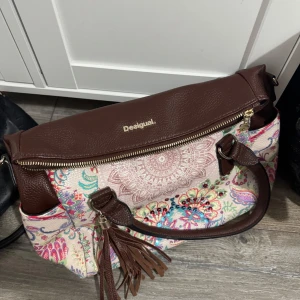 Mönstrad handväska från Desigual - Säljer en unik handväska från Desigual med bruna skinn-detaljer och färgglatt blommigt mönster. Väskan har dragkedja, två handtag och dekorativa tofsar. Perfekt för dig som vill sticka ut med en färgglad accessoar.