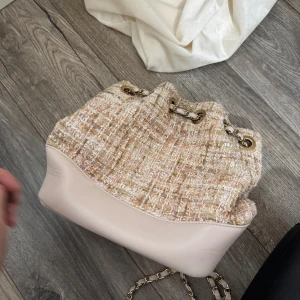 Beige och rosa axelväska med kedja - Trendig axelväska i beige och ljusrosa med grovt vävt tyg upptill och slät botten. Väskan har guldfärgade metalldetaljer och en kedjerem. Perfekt för att lyfta din outfit med en chic touch.