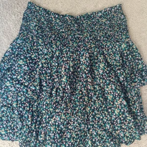 Blommig volangkjol - Supersöt kort blommig kjol från Amisa Collections, köpt på ullared, med volanger och smock i midjan. Mörk botten med små blommor i grönt, rosa, vitt och gult. Perfekt för en somrig look och enkel att matcha med en topp. Superfin och nästintill aldrig använd. 50kr 💕