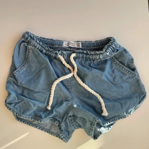 Jeansshorts med snörning från Cubus - Säljer ett par ljusblå jeansshorts i storlek 134/140 från Cubus! Shortsen har elastisk midja och vit, grov snörning framtill. De har två fickor på sidorna och är tillverkade i ett mjukt denimtyg. 