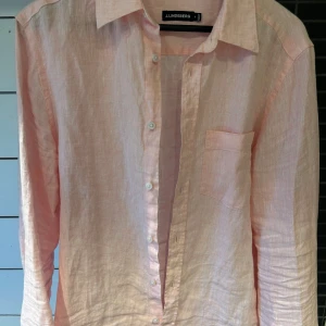 Ljusrosa linneskjorta från J.Lindeberg - Säljer en stilren ljusrosa skjorta från J.Lindeberg i svalt linne. Skjortan har klassisk krage, bröstficka och knäppning framtill. Perfekt för varma dagar när du vill vara både bekväm och snygg. Bara höra av er om det finns några frågor