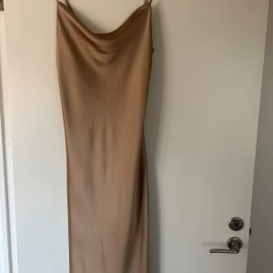 Champagnefärgad långklänning från NA-KD - Säljer en elegant långklänning från NA-KD i champagnefärgad satin. Klänningen har smala axelband och en mjukt draperad halsringning. Perfekt för dig som vill ha en stilren och trendig look.Det är denna klänning (inte rätt färg) :  https://www.na-kd.com/sv/produkter/waterfall-midi-slip-dress-brown-1100-008134-0017  Oanvänd, endast testad