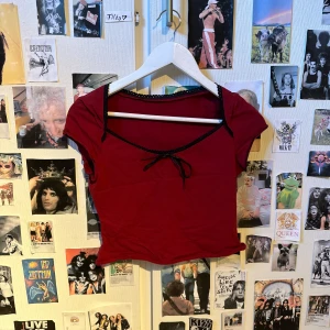 Röd croppad topp med svarta detaljer - Snygg röd croppad topp med svarta band och spetsdetaljer vid halsringningen. Toppen har korta ärmar och en söt knytning framtill. Perfekt för dig som vill ha en trendig och ungdomlig stil.