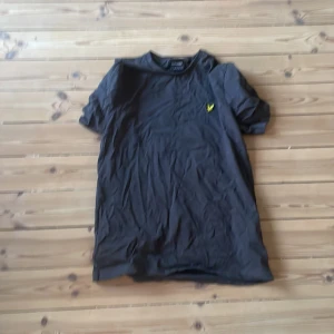 Mörkgrå t-shirt från Lyle & Scott - Mörkgrå t-shirt från Lyle & Scott med klassisk rund hals och korta ärmar. På bröstet finns en broderad gul logotyp i form av en fågel. Perfekt för en stilren och enkel look. Storlek M i mäns. Helt ny men blev för stor för mig. Skriv vid funderingar!
