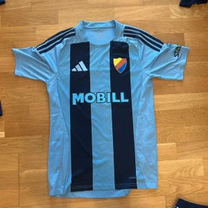 Djurgårdens IF fotbollströja med nummer 11 - Säljer en ljusblå och mörkblå randig Djurgårdens IF fotbollströja från Adidas med klubbmärke, sponsortryck och nummer 11 på ryggen. Tröjan har korta ärmar och klassiska Adidas-ränder på axlarna.