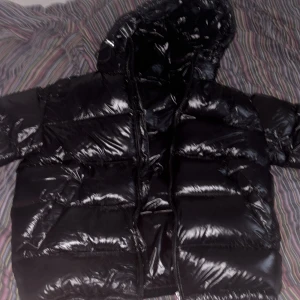 Svart pufferjacka från ZARA med huva - Säljer en svart pufferjacka från ZARA med glansig finish och huva. Jackan har dragkedja framtill och stora fickor. Perfekt för kyliga dagar när du vill hålla dig varm och stilsäker.