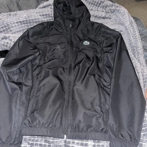 Svart vindjacka från Lacoste med huva - Svart vindjacka från Lacoste med huva och dragkedja framtill. Storlek S, knappt använd.