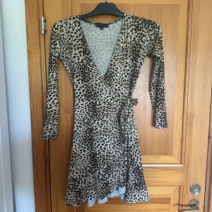 Leopardmönstrad omlottklänning - Snygg leopardmönstrad omlottklänning med lång ärm och v-ringning. Klänningen har knyt i midjan och är i en kort modell. Perfekt för dig som vill sticka ut med ett djurmönster.