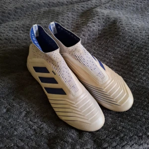 Adidas fotbollsskor med blå ränder - Säljer ett par vita Adidas fotbollsskor med blå detaljer och tre klassiska ränder på sidan. Skorna har en strumpliknande passform utan snörning och är designade för spel på gräs. Perfekta för dig som vill ha stilrena och moderna fotbollsskor.