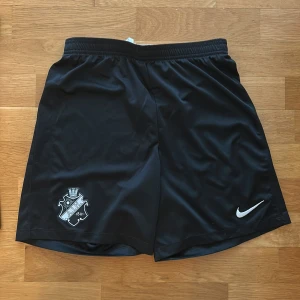 Svarta träningsshorts från Nike med AIK-logga - Svarta träningsshorts från Nike med AIK:s klubbmärke på ena benet och Nike-logga på det andra. Tillverkade i Dri-Fit-material som andas och har resår i midjan med snörning för bästa passform.