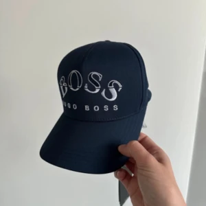 Mörkblå keps från Hugo Boss - Snygg mörkblå keps från Hugo Boss med stor vit logotyp framtill. Klassisk böjd skärm och justerbar passform baktill. Perfekt accessoar för en sportig och stilren look.