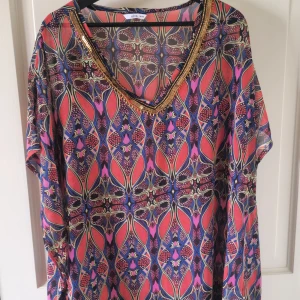 Mönstrad klänning med paljettkant från ellos - Färgglad klänning från ellos med abstrakt mönster i rosa, blått och lila. V-ringad hals med dekorativ paljettkant. Lätt och luftig modell med korta ärmar, perfekt för varma dagar.