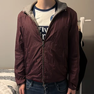 Moncler windbreaker  - Säljer en vinröd windbreaker från Moncler med huva och grått innerfoder. Jackan har dragkedja framtill och Moncler-logga på ärmen. Perfekt för blåsiga dagar och enkel att matcha med jeans eller träningskläder. Modellen är 190cm, sitter som L