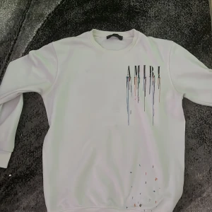Vit sweatshirt från Amiri med färgstänk - Säljer en vit sweatshirt från Amiri med tryckt logga och färgstänk i flera färger både fram och bak. Tröjan har rund hals och långa ärmar. Perfekt för dig som gillar streetwear med unik design.