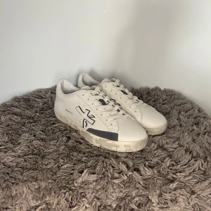 Premiata Sneaker - HELT NYA! Säljer ett par stilrena vita sneakers från Premiata med grå detaljer och snörning. Skorna har en platt sula och rund tå, samt märkets logga på sidan. Perfekta för dig som gillar klassisk och enkel design. Dustbag ingår. 