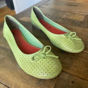 Ljusgröna ballerinaskor med rosett fån Tamaris - Snygga ljusgröna ballerinaskor från Tamaris med små hål i mönstret och en söt rosett framtill. Skorna har rund tå och platt sula, samt röd innersula som kontrast. För små,så toppenskick.