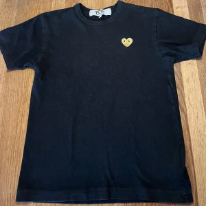 Svart t-shirt från Comme des Garçons Play - Svart t-shirt från Comme des Garçons Play med ett litet broderat gult hjärta med ögon på bröstet. Perfekt för sommaren, har tyvärr ett pyttelitet hål nära etiketten.