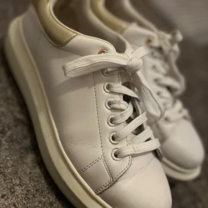 Vita sneakers med gulddetalj - Snygga vita sneakers med metallicguld detalj vid hälen och vita snören. Skorna har en tjock, platt sula och rund tå. Perfekta för dig som vill ha en stilren men ändå trendig look.