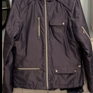 Grafit grå  jacka från Mountain Horse - Säljer en grafit grå  jacka från Mountain Horse med dragkedjor och flera fickor framtill. Jackan har lång ärm, quiltat innerfoder och broderad logga på insidan. Perfekt för dig som gillar stilren och funktionell design.