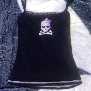 Svart döskalle linne med spets - Svart linne med smala axelband och spetskant upptill. På bröstet finns en broderad döskalle  med rosa rosett och vita detaljer. Sömmen längst ner har en kontrasterande vit dekorsöm som ger en extra touch.