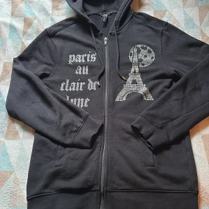 Svart hoodie med Eiffeltorn - Svart hoodie från BINGLUCID med dragkedja, huva och snörning. Framsidan har coola strasstenar som bildar Eiffeltornet, måne och texten 'paris au clair de lune'. Mjuk bomullskänsla och långa ärmar. Perfekt för dig som gillar statement-plagg med Paris-vibe.