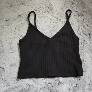 Svart ribbad linnetopp - Ett svart croppad linne med smala axelband och v-ringning. Toppen är ribbad. Har några trådar på axelbandet men det märks inte när man använder det. Skriv om du har funderingar.