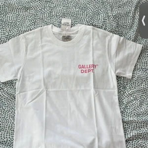 Vit t-shirt från Gallery Dept. - Vit t-shirt från Gallery Dept. med rosa tryck fram och stort tryck på ryggen med texten 'GALLERY DEPT. HOLLYWOOD, CA.'. Klassisk passform och rund halsringning. Perfekt för dig som gillar streetwear och enkel stil.