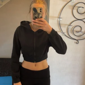 Svart croppad zip-up - Svart croppad zip-up hoodie med luva och dragkedja framtill. På ryggen står det 'Angel' i stora bokstäver av glittrande strass. Tröjan har långa ärmar och en tight passform som sitter snyggt på kroppen. Slutar strax över naveln. Storlek M men jag skulle snare säga XS/S!☺️