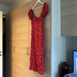 Röd blommig långklänning med puffärm - Superfin röd långklänning med små vita blommor och puffiga korta ärmar. Klänningen har en söt v-ringning och smockad överdel som ger en romantisk vibe. Dragkedja baktill. Använd 1 gång!