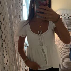 Vit blus med puffärm och knyt - Supersöt vit blus från Gina med puffiga korta ärmar och två knyt framtill. Blusen har en lätt rynkad struktur och volang nedtill som ger en romantisk vibe. Perfekt till jeans eller kjol för en fräsch look.
