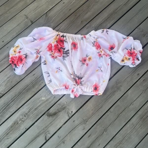 Blommig offshoulder blus med puffärm - Superfin blommig blus i vitt med röda och gula blommor. Offshoulder-modell med stora puffärmar och knytning framtill. Croppad passform och lätt, luftig känsla. Perfekt för en somrig look!