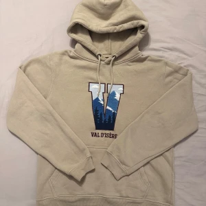 Beige hoodie Val d'Isère S - Beige hoodie från Val d'Isère med stor broderad V på bröstet i blått och vitt med bergmotiv. Tröjan har huva med snörning, känguruficka och är tillverkad i mjuk bomull och polyester. Har endast sålts på Intersport val d’Isere och endast under säsongen 2021/22. Stor i storleken, närmare M/L