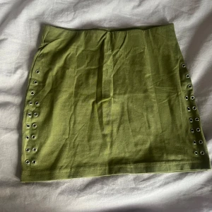 Olivgrön kortkjol med nitar, strl 34 - Snygg olivgrön kortkjol med coola metallöljetter längs sidorna. Kjolen är i stretchig bomullsmix och har en tight passform som sitter snyggt på kroppen. Perfekt för dig som vill ha en edgy och trendig look.