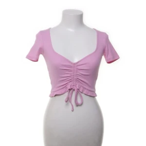 Rosa croppad topp med snörning från Zara - Supersöt ljusrosa croppad topp från Zara med korta ärmar och v-ringning. Toppen har ribbad struktur och snygg snörning framtill som ger en rynkad effekt. Perfekt för varma dagar och ger en trendig vibe till din outfit.