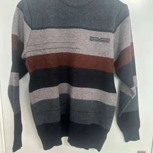 Randig stickad tröja i mörka toner - Snygg stickad tröja med breda ränder i svart, grått, brunt och mörkblått. Tröjan har rund halsringning och långa ärmar. Perfekt för dig som gillar en chill och stilren look med lite retrovibbar.