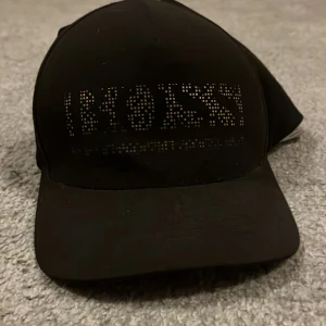 Svart keps från Hugo Boss - Snygg svart keps från Hugo Boss med coolt perforerat BOSS-tryck framtill. Justerbar rem baktill med metallspänne där BOSS-loggan är ingraverad. Klassisk böjd skärm och stilren design, perfekt för dig som gillar streetwear och premiumkänsla.