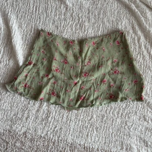 Grön blommig kortkjol med dragkedja - Supersöt ljusgrön kortkjol med rosa blommor och diskreta bruna blad. Kjolen har en smickrande A-linjeform och stängs med en dragkedja i sidan. Perfekt för vår och sommar, till sneakers eller sandaler. Materialet känns lätt och luftigt.