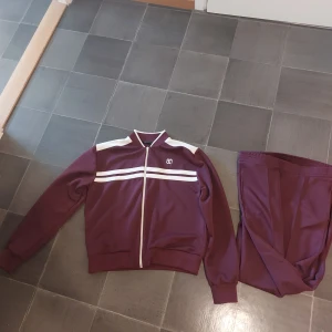 Vinröda mjukisbyxor med matchande jacka - Säljer ett par vinröda mjukisbyxor från Zara med matchande jacka. Byxorna är i mjukt material och har en sportig look. Perfekta för chill eller träning. Snygg vit detalj på jackan och klassisk passform.Andvänd en gång.