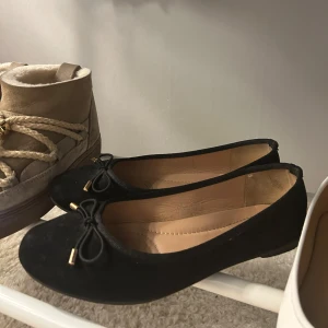 Svarta ballerina - Stilrena svarta ballerinaskor med rund tå och söt rosettdetalj framtill. Skorna har en klassisk, låg profil och är perfekta för en enkel men chic look. Insidan är ljusbeige och materialet känns mjukt och bekvämt.