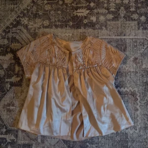 Champagnefärgad kofta i satin - Supersöt blus i champagnefärgad satin med korta ärmar och plisserade detaljer upptill. Den har en lös passform och ett glansigt material som ger en lyxig känsla. Perfekt för dig som gillar romantiska och trendiga plagg.