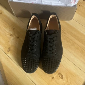 Svarta Louboutin sneakers med nitar - Snygga svarta sneakers från Christian Louboutin med klassisk röd sula och svarta nitar på tån. Skorna har snörning, rund tå och är tillverkade i mocka med detaljer i skinn. Perfekta för dig som vill sticka ut med en edgy och lyxig stil.