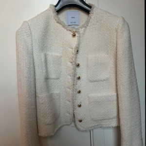 Vit cardigan jacka från MANGO - Snygg vit cardigan jacka från MANGO med guldknappar och fyra fickor framtill. Jackan har en strukturerad, lite ruffig yta och är croppad i modellen. Perfekt för dig som vill ha en chic och trendig look.