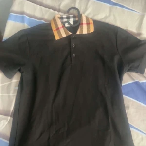 Svart Burberry t-shirt  - Svart t-shirt från Burberry med klassisk rutig krage i beige, svart, vitt och rött. Modellen har korta ärmar och tre knappar framtill. Snygg och stilren design som sticker ut med den ikoniska Burberry-kragen.