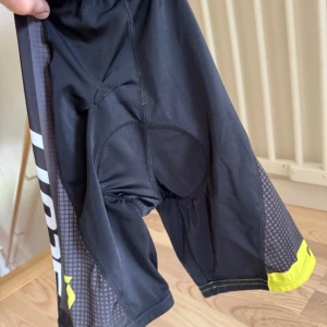 Scott Svarta cykelshorts med vaddering - Säljer ett par svarta cykelshorts från Scott med vaddering för extra komfort. Shortsen har gula detaljer och tryck på sidan samt elastisk midja. Perfekta för dig som gillar att cykla längre sträckor och vill ha en sportig look.