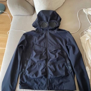C.P. Company Goggle Jacket navy - Säljer en mörkblå Goggle Jacket från C.P. Company med ikoniska glasögon i huvan. Jackan har flera fickor framtill, dragkedja och är tillverkad i stretchigt soft shell-material av återvunnen nylon. Perfekt för dig som vill sticka ut med en unik streetwear-look.